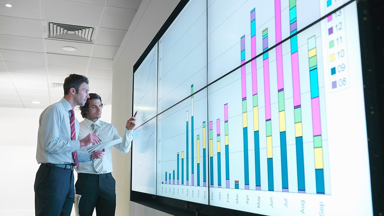 Business men analyse reports displayed on a large digital screen