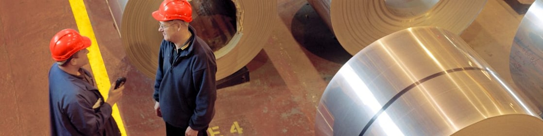 Two factory workers standing next to rolls of steel