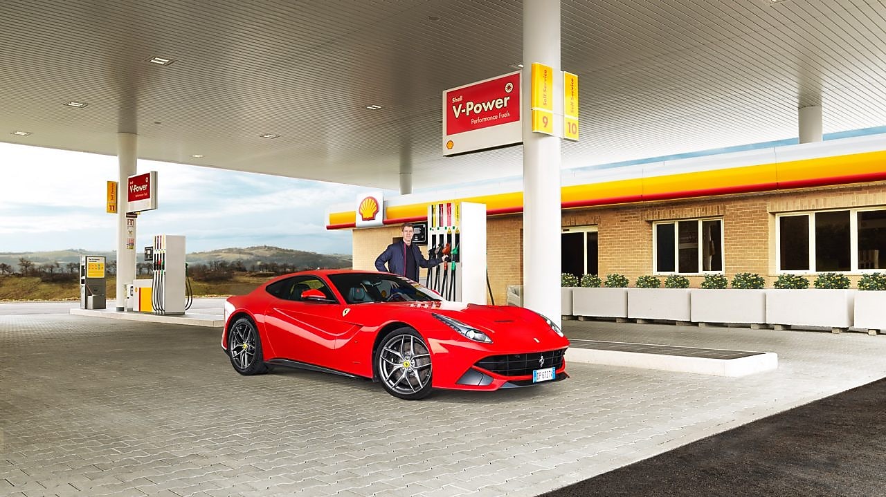 A red Ferrari sitting on a Shell station forecourt with a man leaning on a petrol pump